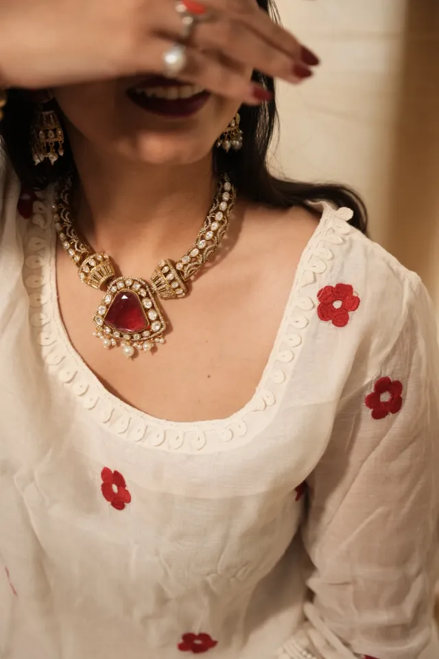 A woman in a white kurta wearing a red-and-gold necklace and jhumkas